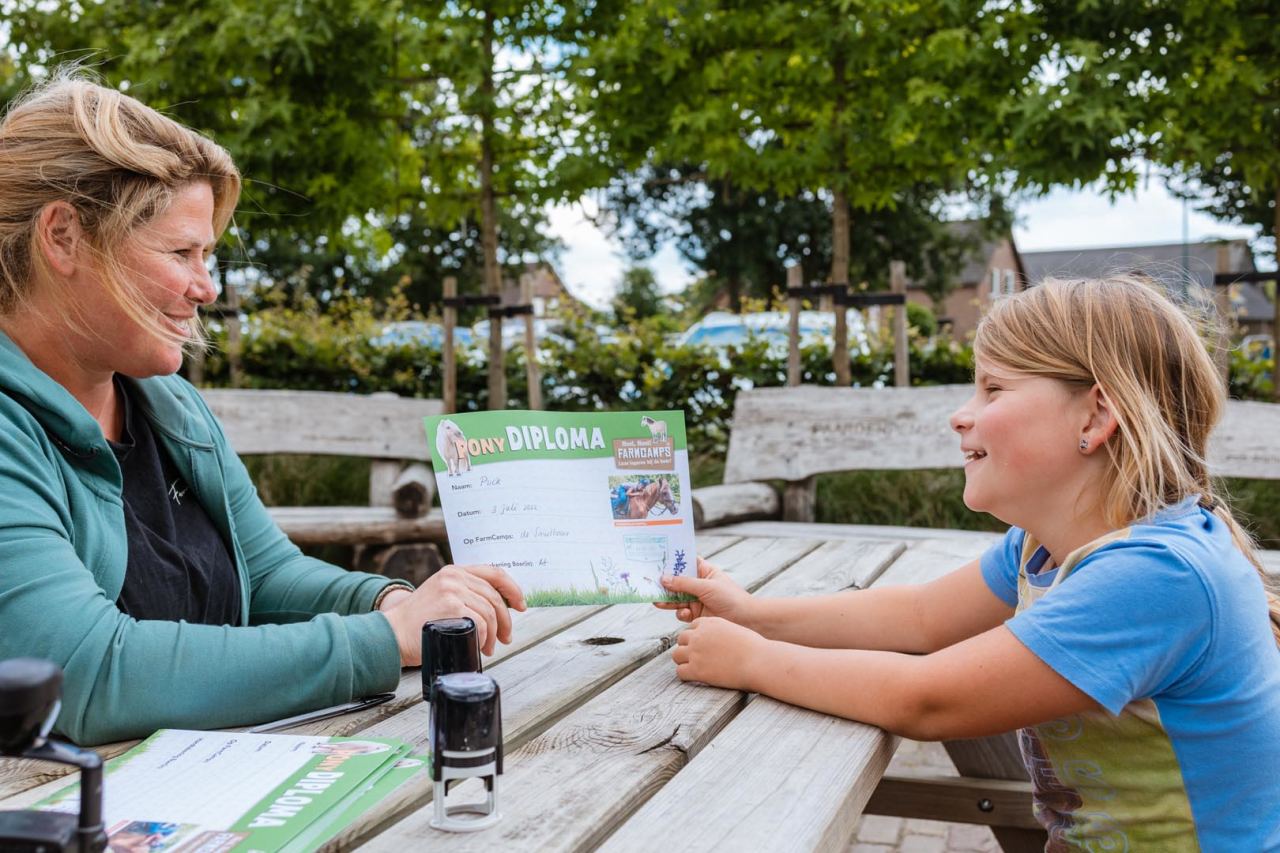 Kind ontvangt trots een FarmCamps Pony Diploma van de boerin aan een picknicktafel op de boerderij