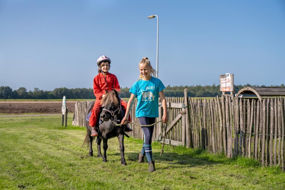 Kind maakt een ponyrit op de boerderij terwijl een ander kind de pony begeleidt bij FarmCamps