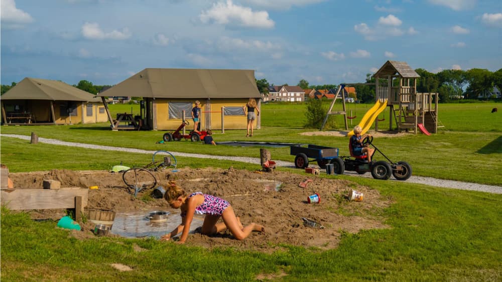 Kinderen spelen in de zandbak en rijden op skelters bij de tenten van FarmCamps De Oostermaat