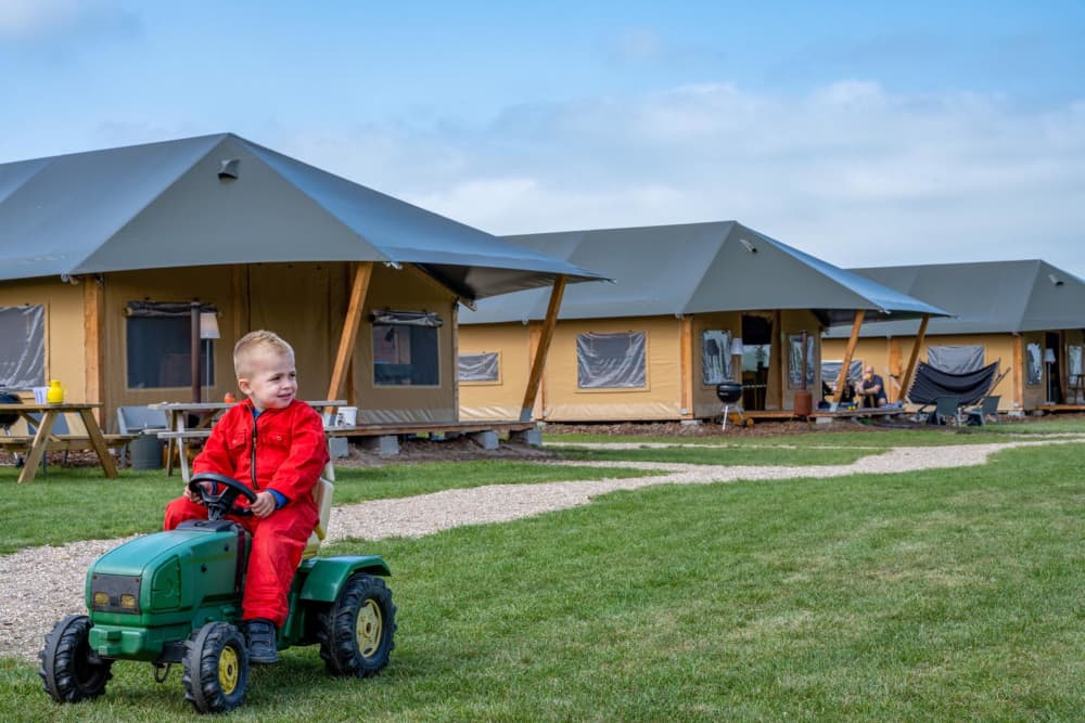 Kind fährt mit einem Spielzeugtraktor vor den Luxuszelten während eines Urlaubs auf dem Bauernhof bei FarmCamps