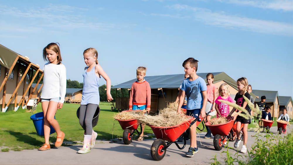Kinder laufen mit einer Schubkarre voller Heu über den Bauernhof während eines Urlaubs bei FarmCamps