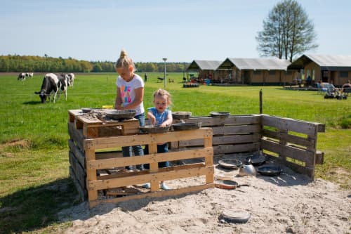 Children playing happily in front of the glamping tents at FarmCamps 't Looveld, with wide open fields and grazing cows in the background.